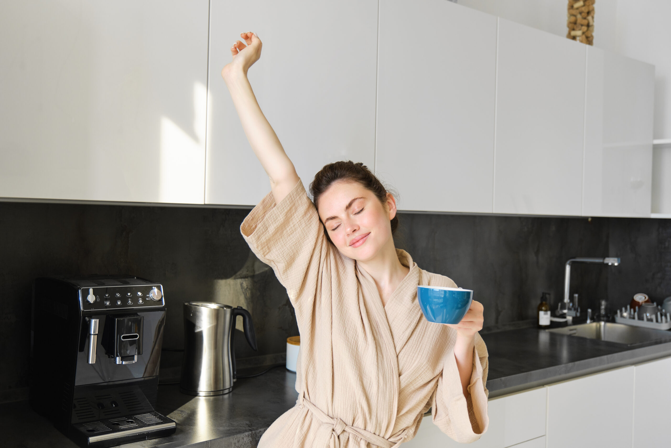 Portrait of happy girl dancing with coffee in the kitchen, wearing bathrobe, enjoying her morning routine.